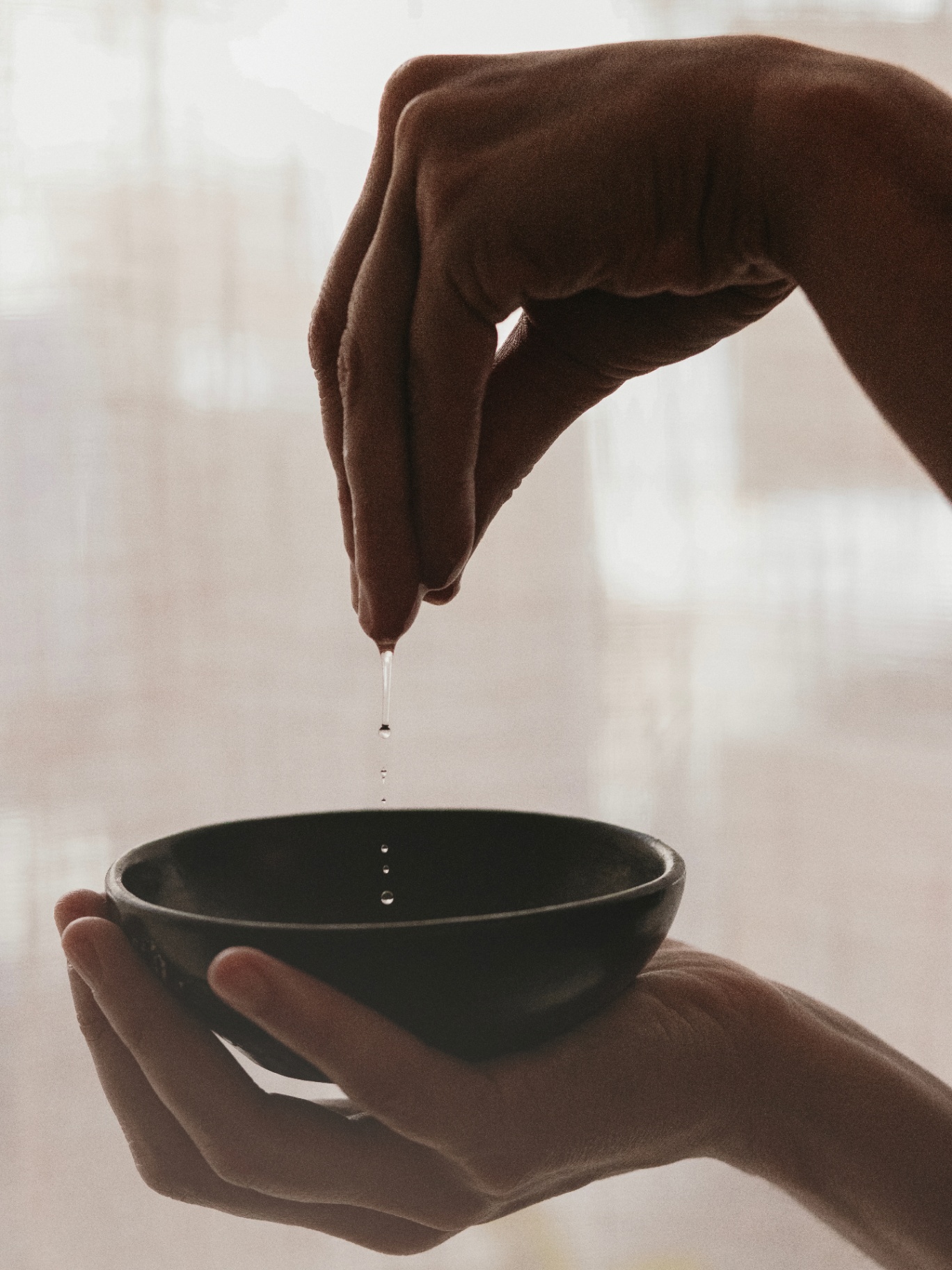 Warm oil poured between two cupped hands into a small dark bowl
